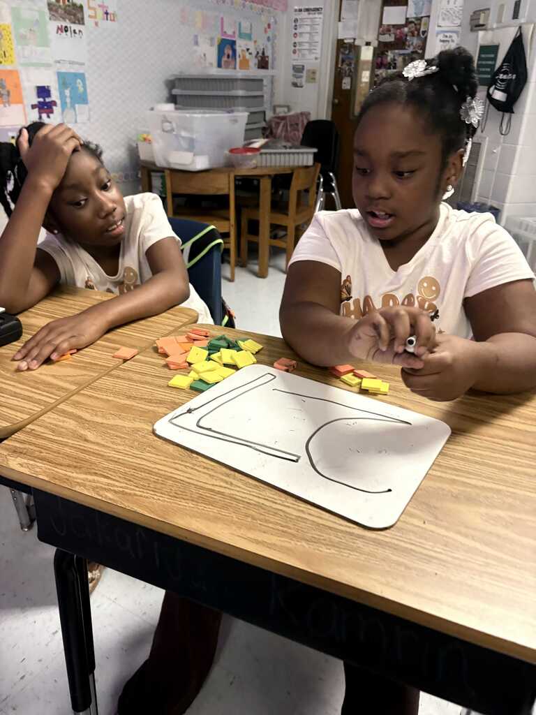 Two girls sit at a table, one focused on colored shapes on a board, the other looking at them.