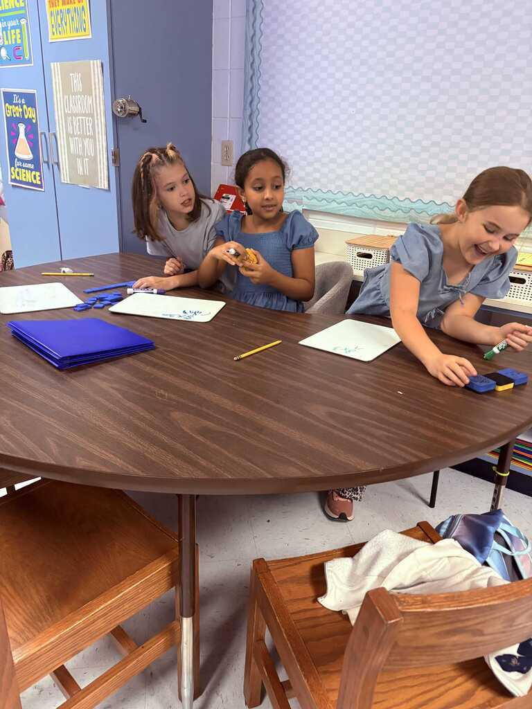 Three children seated at a round table. Each child is holding an object and smiling. Papers, pens, and a blue binder on the table.