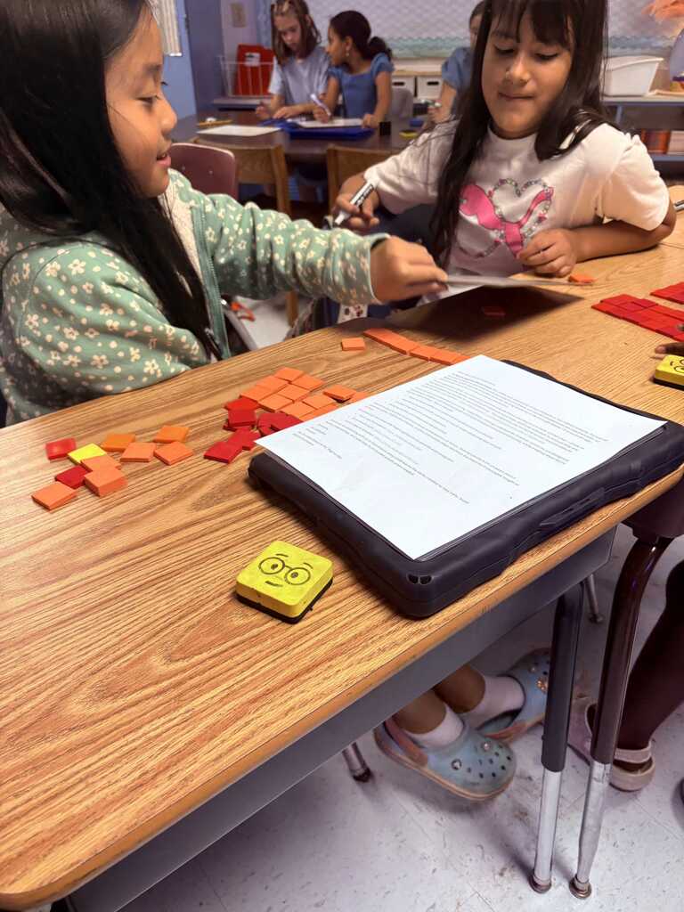 Two young children are seated at a table, arranging colorful tiles. A tablet and a binder with papers are on the table.