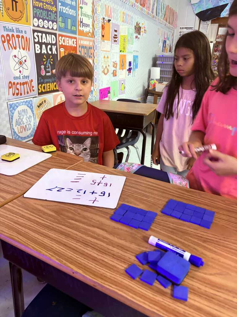 Three children in a classroom, one boy in red and two girls, one in pink, work at a table with blue shapes and paper.