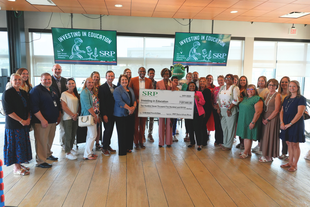 A group of adults stands in a room holding a large check and banners that read "Investing in Education."