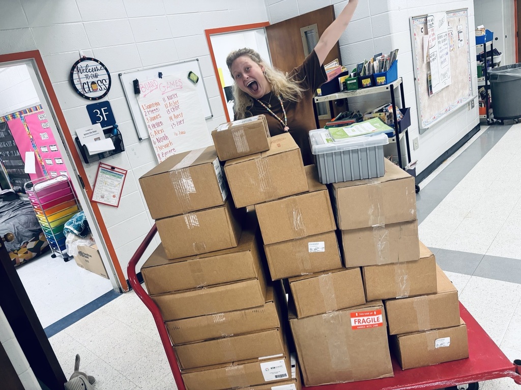 A woman pushes a red cart with many cardboard boxes inside a room with white walls.
