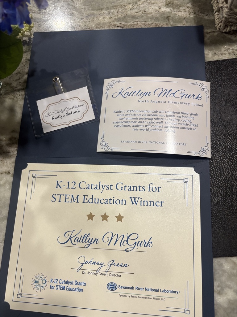 A certificate and badge sit on a blue folder. The certificate reads "K-12 Catalyst Grants for STEM Education Winner".