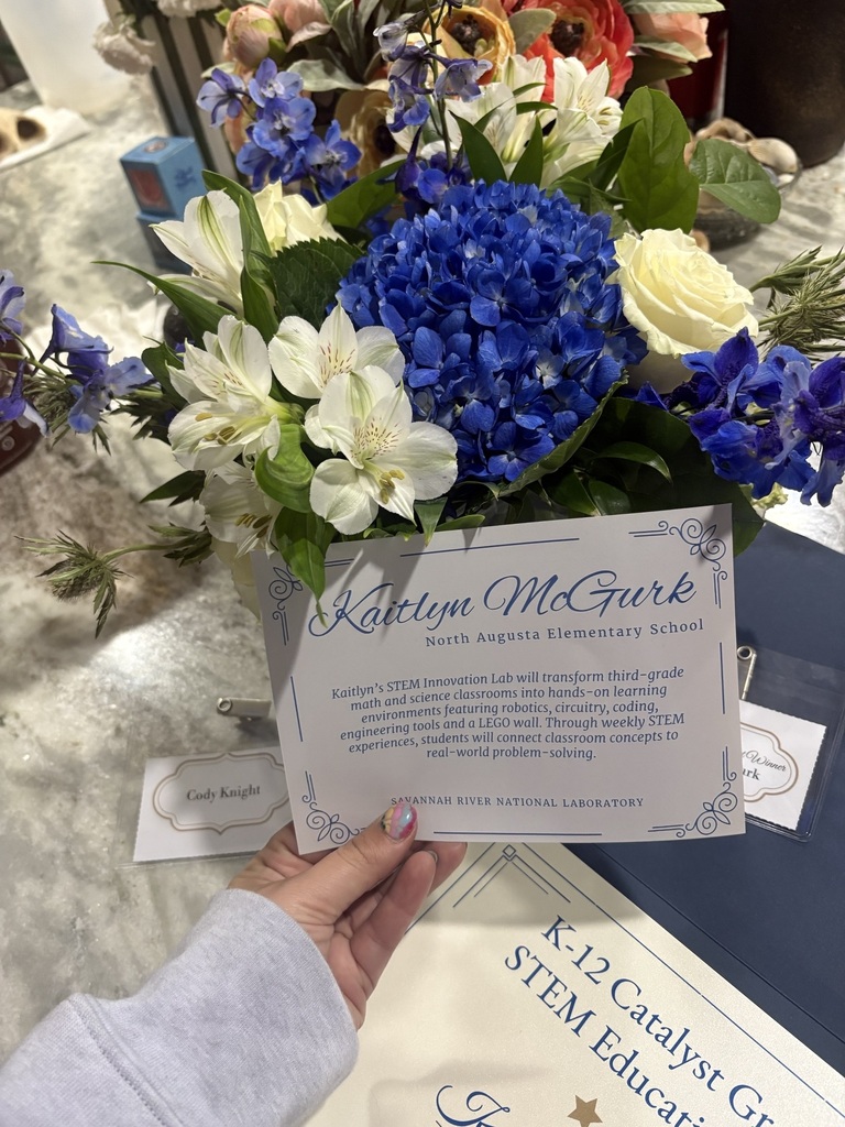 A person holds a certificate with blue flowers in the background. A blue table with white flowers and a certificate titled "STEM Educator" is visible.