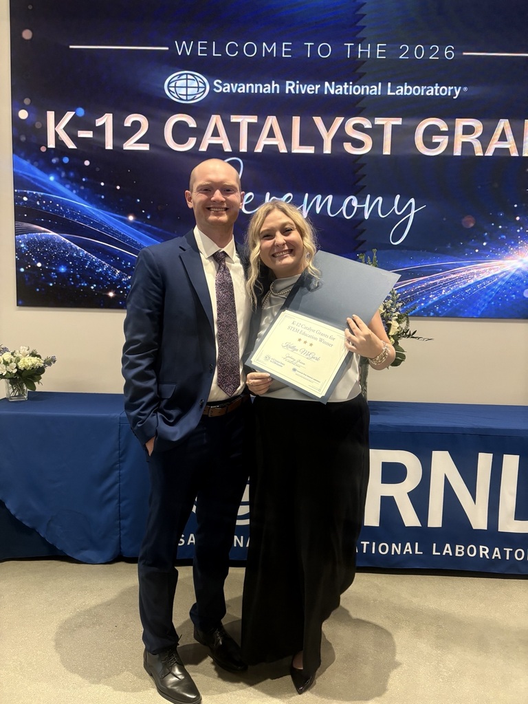 A man and a woman in formal wear pose for a photo while holding a certificate. Behind them is a blue table with a white banner reading "K-12 Catalyst Grand".