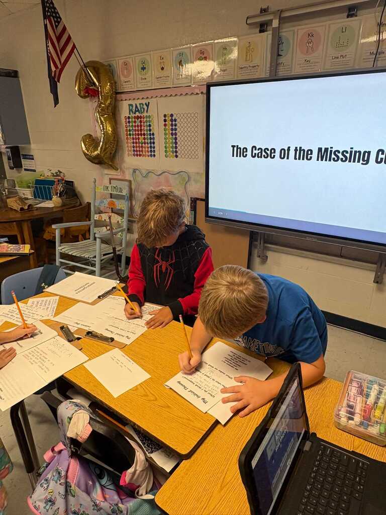 Children sitting at a desk with laptops and papers, one writing, one looking at a screen. A large screen in the background shows "The Case of the Missing C".