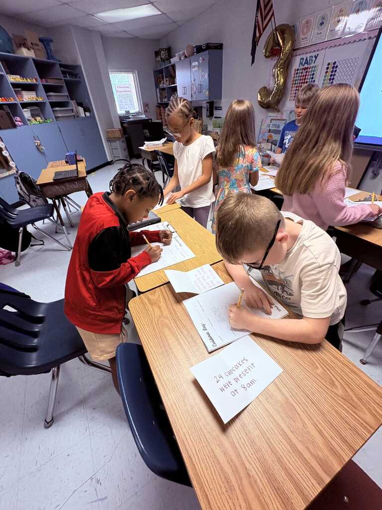 A classroom with desks where several young students work on papers. One boy wears glasses.