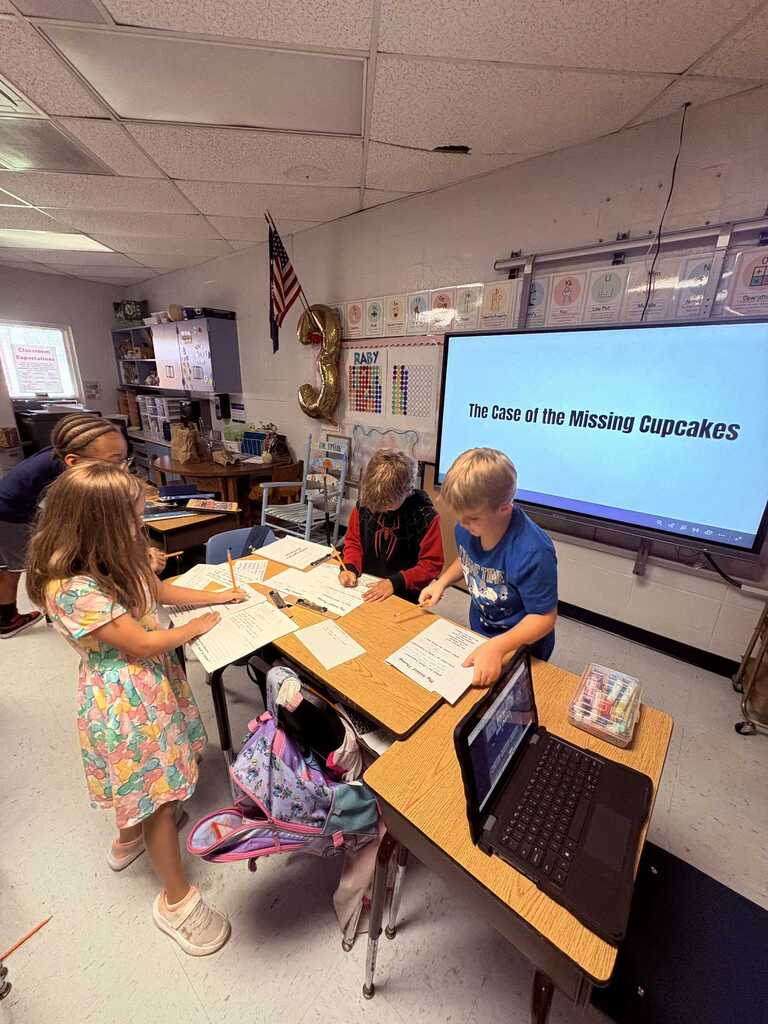 Children in a classroom with a projector screen. They are seated around a table, writing.