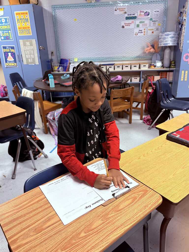 Child sitting at a desk, writing on a notepad with a pencil. Behind, a classroom with desks and chairs.