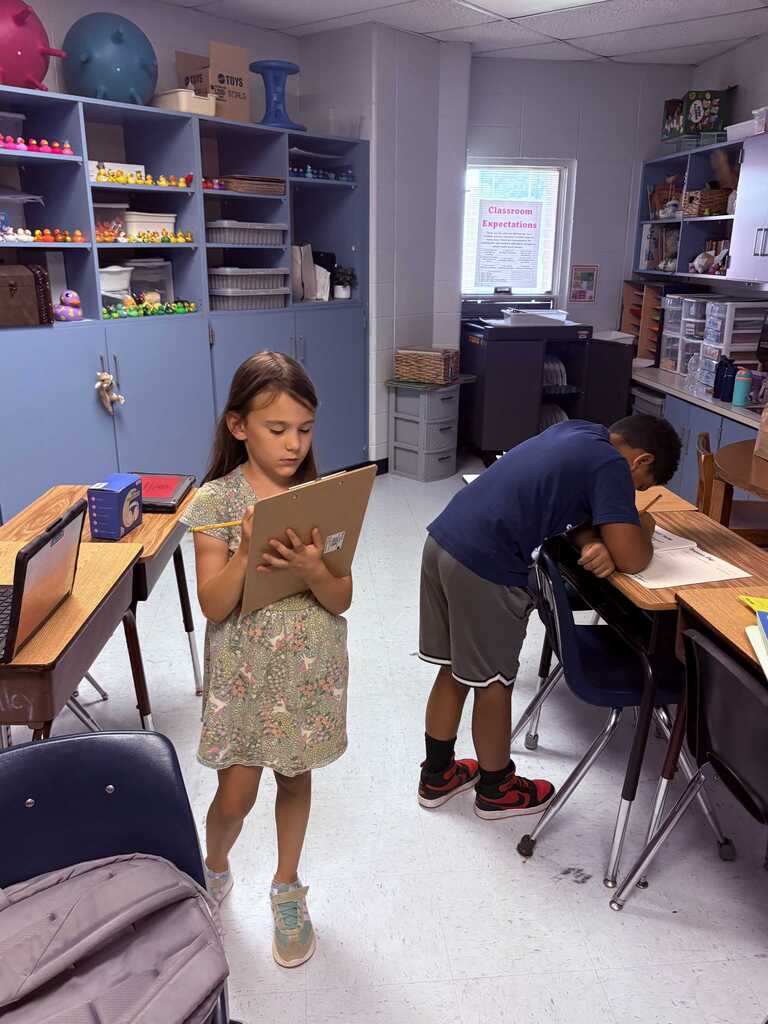 A classroom with a student standing, holding a drawing board, and another student seated, writing at a desk.