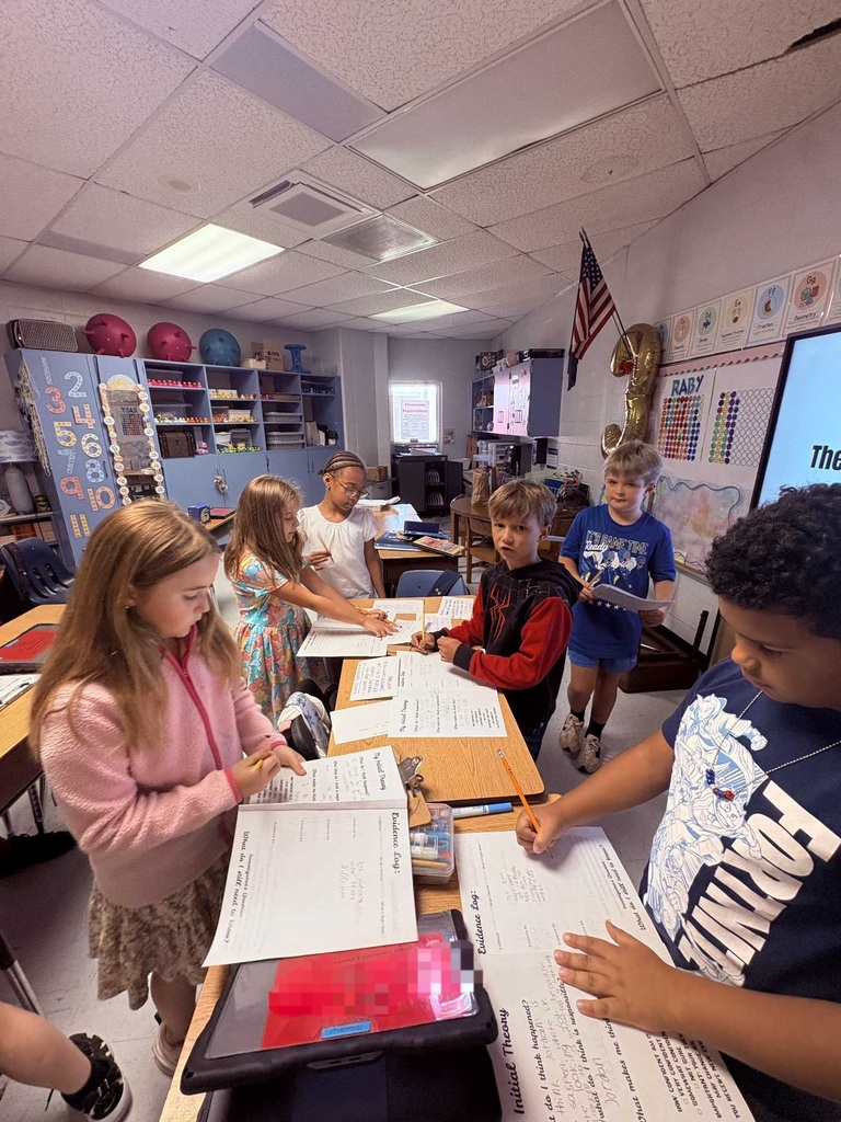 A group of children in a classroom working on papers. A screen displays "The". A flag and books are on the wall.