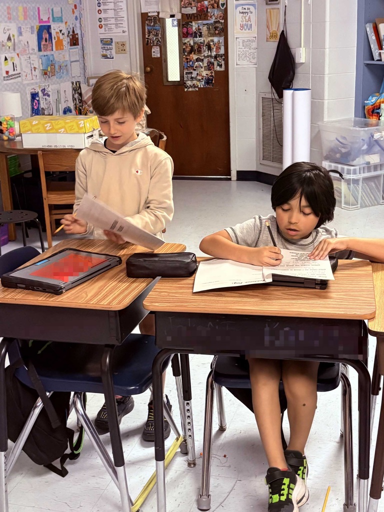 Three children in a classroom sit at desks with books and a tablet, focusing on their work.