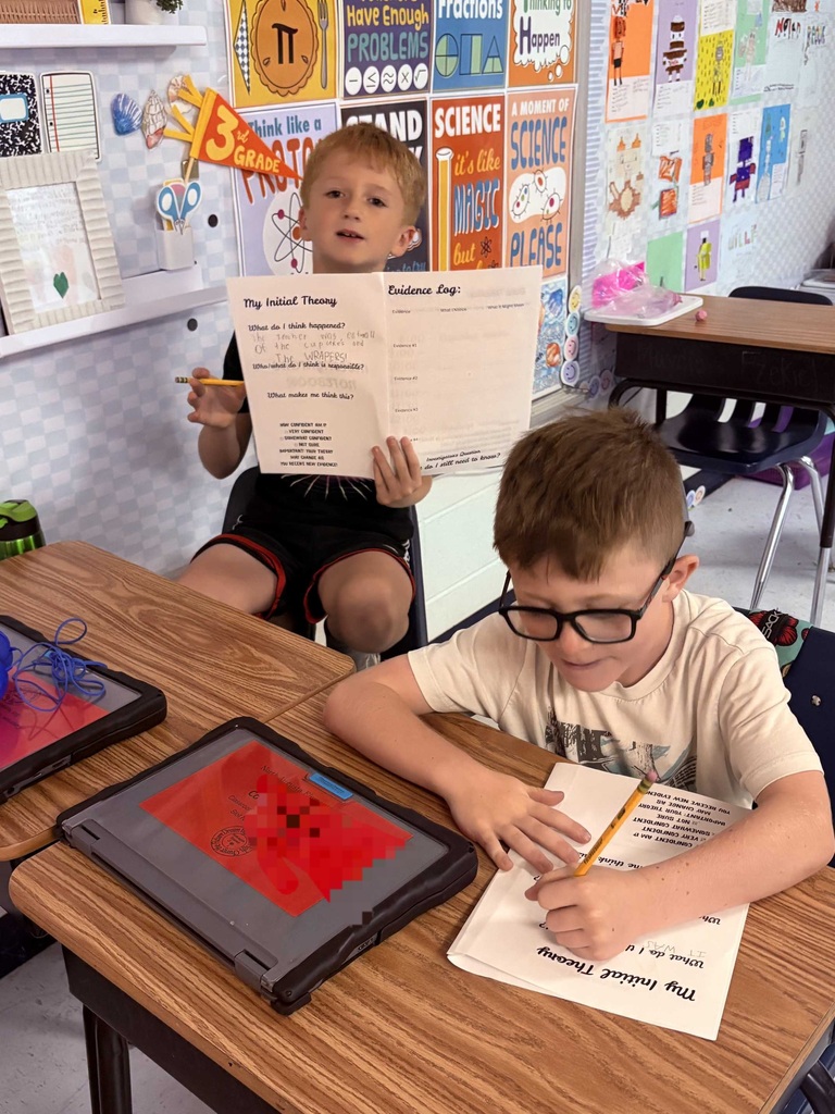 Two children, one with glasses, seated at desks. One holds a paper with writing and a pencil, the other holds a tablet.