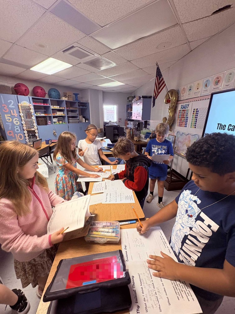 Group of children at tables in a classroom. One child holds a tablet. A projector screen is on the wall behind them.