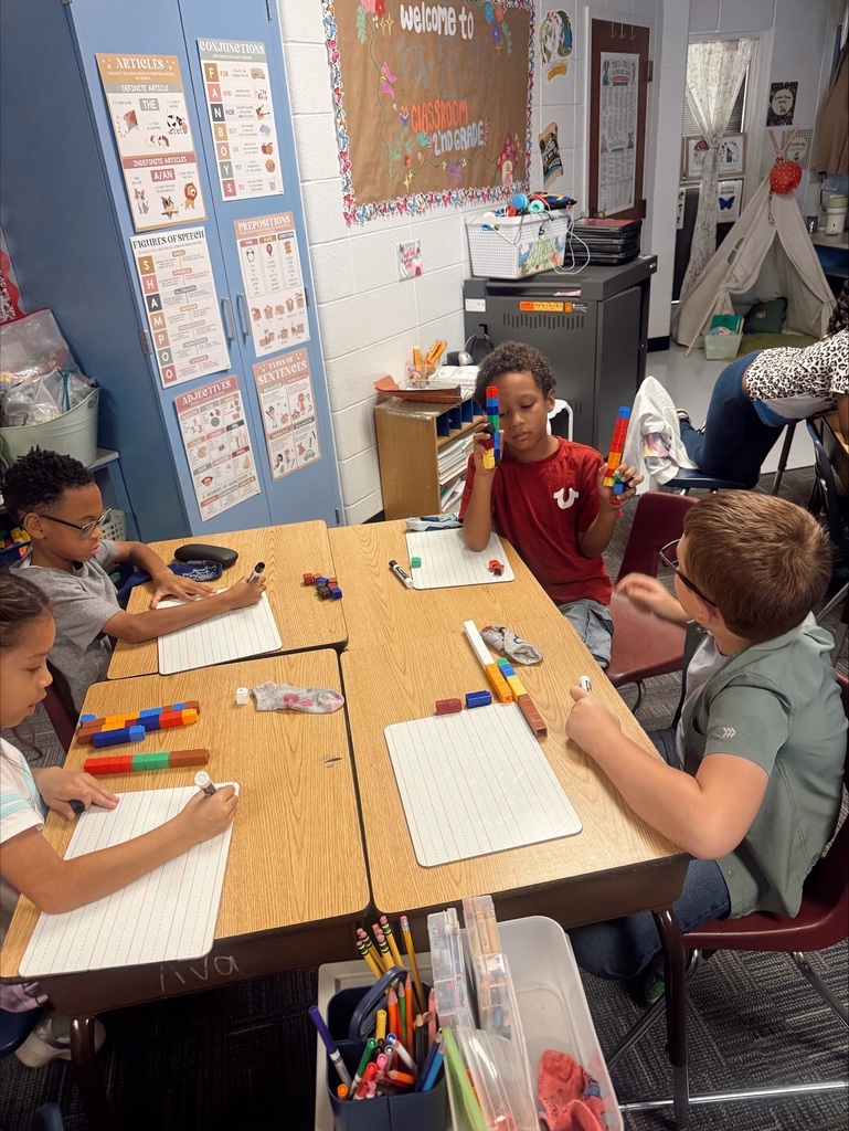 Four children seated around a table in a classroom, using colorful blocks. A teacher is visible in the background.