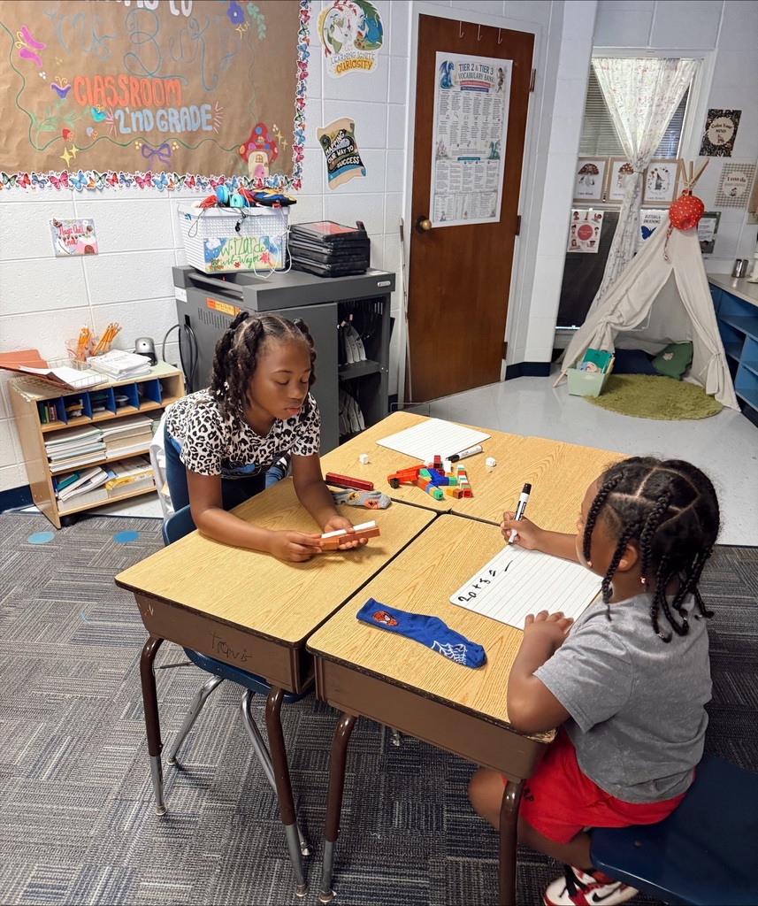 Two children sit at a table, one with a pen and paper, the other with building blocks.