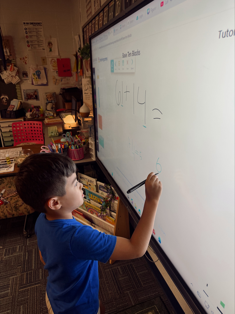 A boy in a blue shirt writes numbers on a whiteboard in a classroom. Behind him are shelves with books.