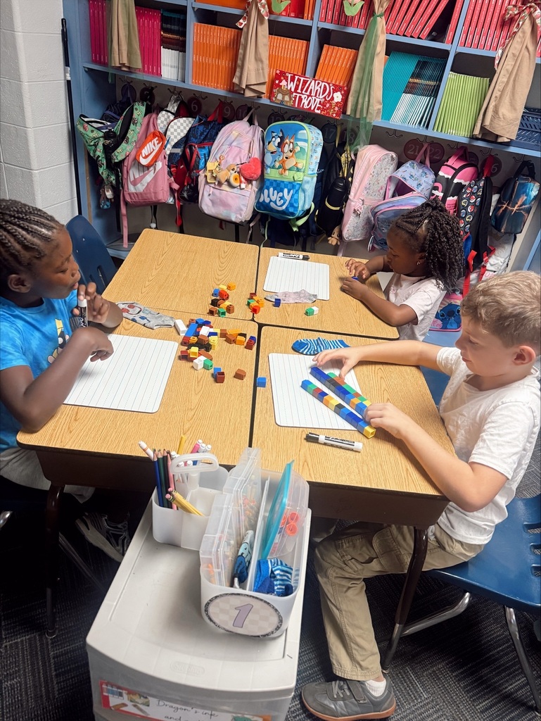 Three children in a classroom, seated around a desk with toys, pencils, and markers. One desk holds a calendar and a pencil holder.