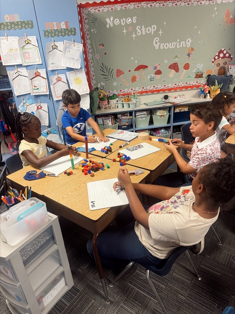 A group of children in a classroom. One child is seated while others sit around a table.