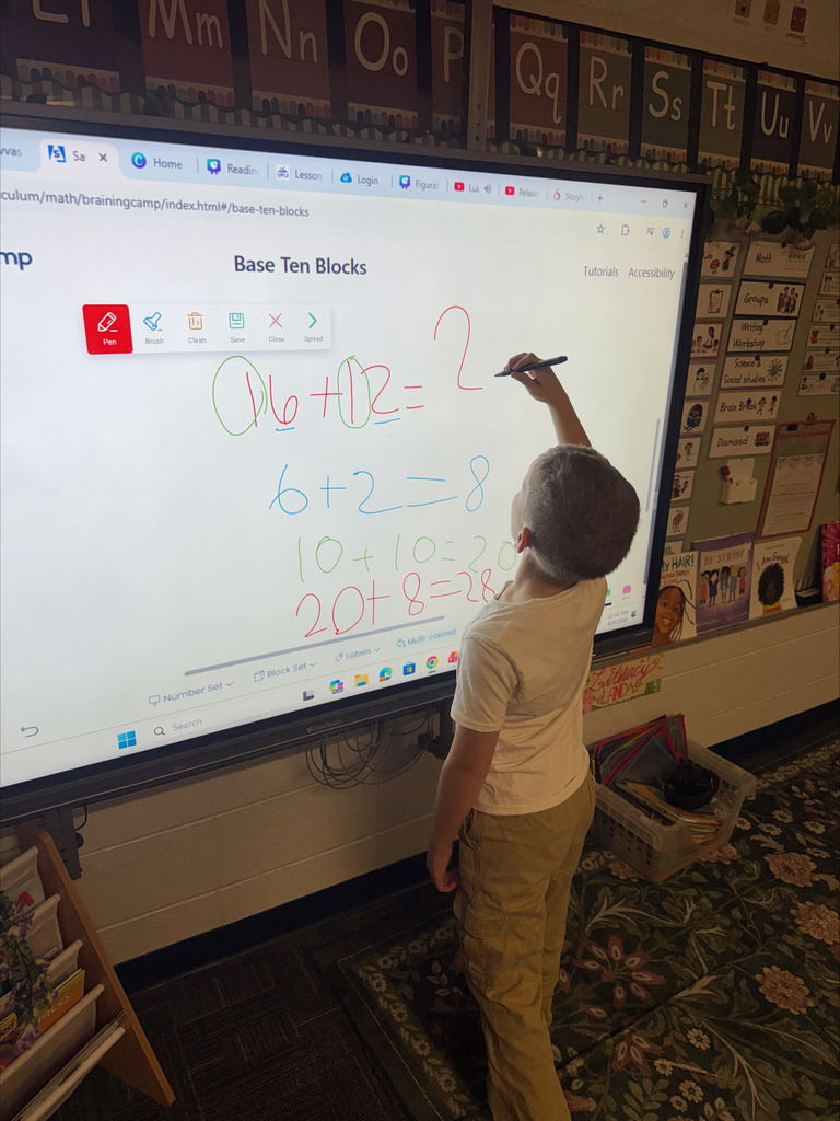 A child writes on a whiteboard, showing math problems and Base Ten Blocks, in a classroom.