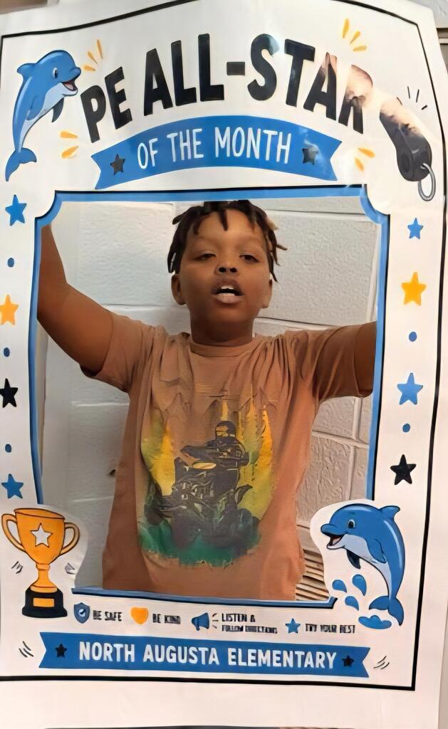 A boy wearing a brown T-shirt stands in a photo frame with blue stars. The frame reads "PE All-Star of the Month."