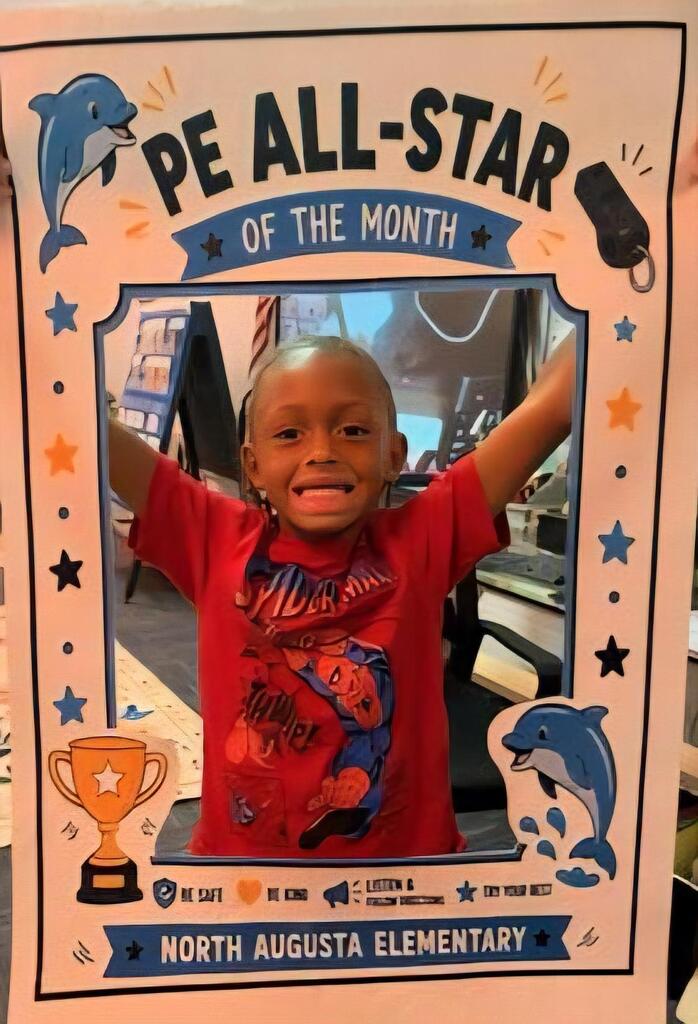 A child smiling and raising arms in front of a photo frame with "PE All-Star of the Month" text.