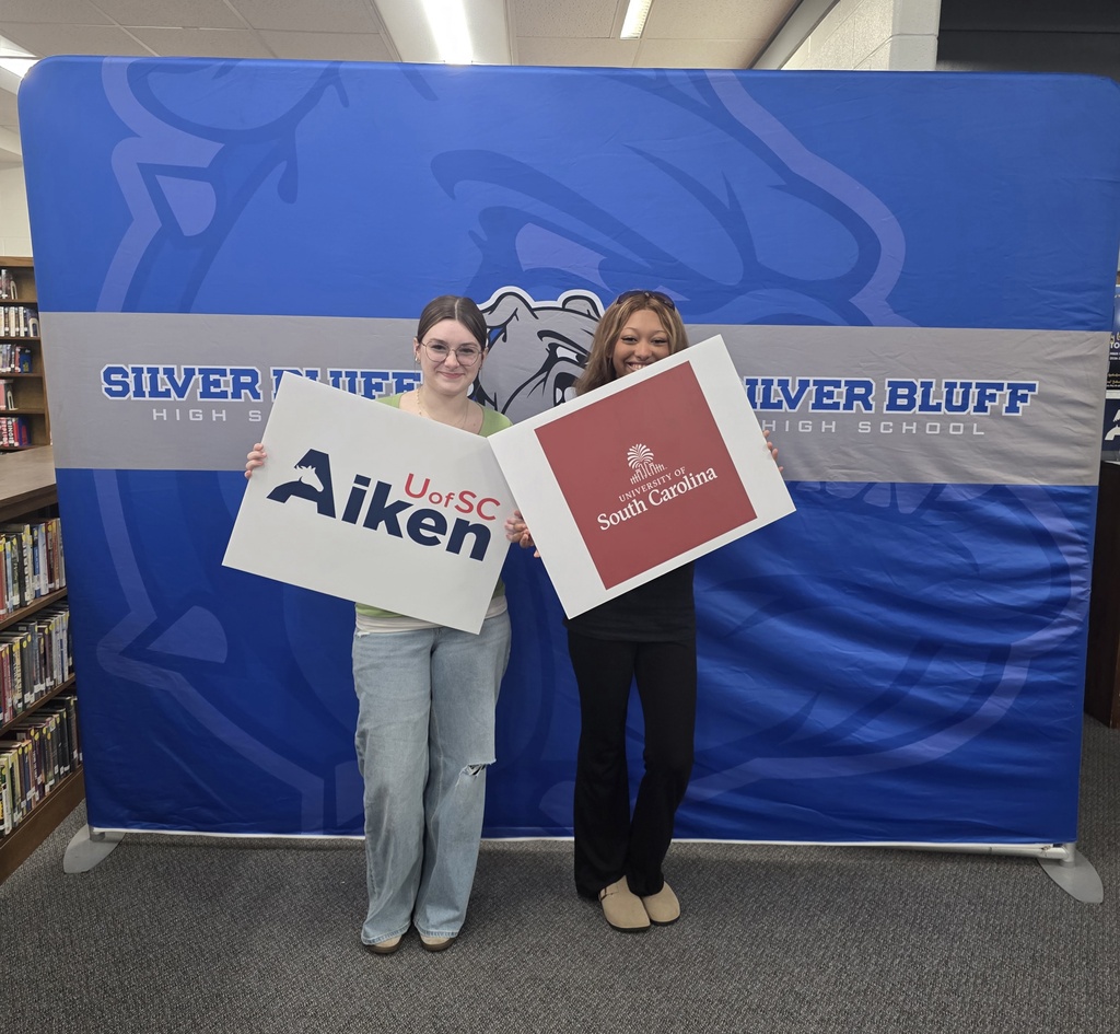 Student in front of Silver Bluff High School blue backdrop holding a college signs