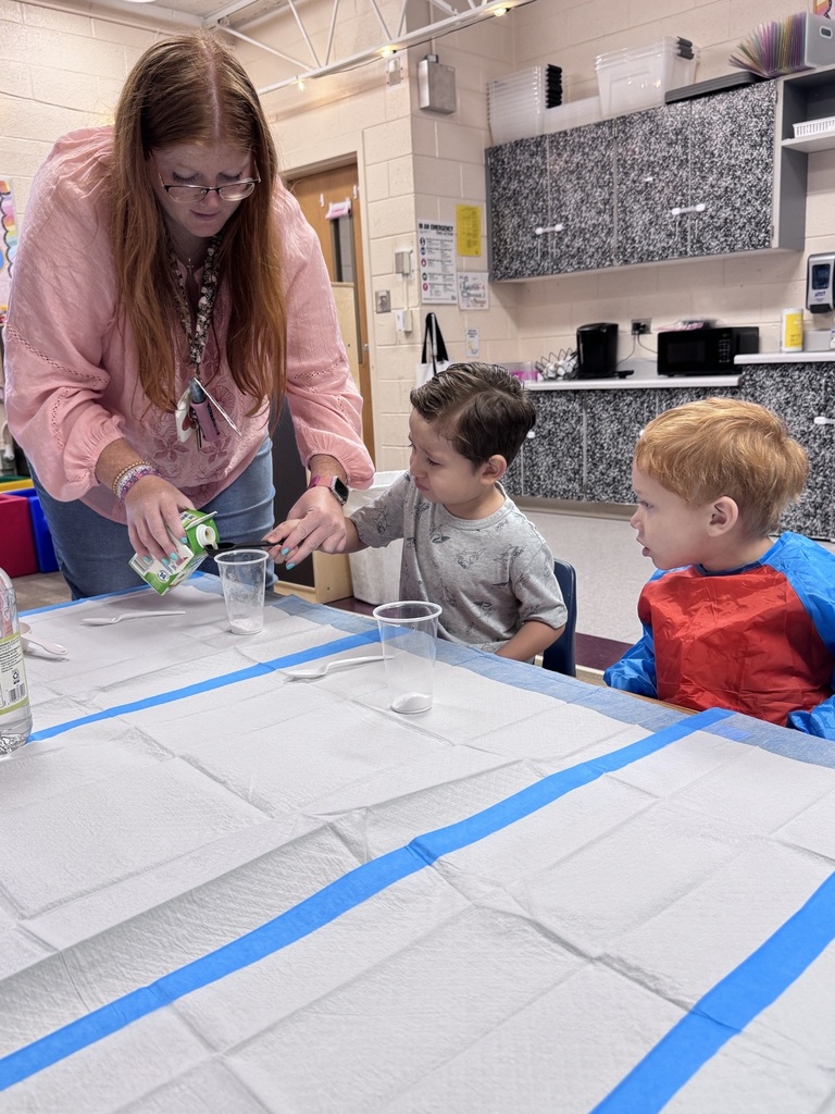 Two boys sit with a cup in front of them while a teacher helps one hold a measuring spoon.