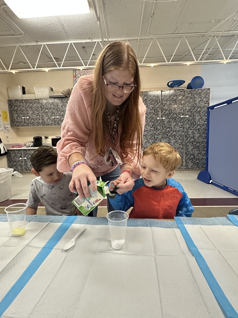 Two boys sit with a cup in front of them while a teacher helps one hold a measuring spoon.