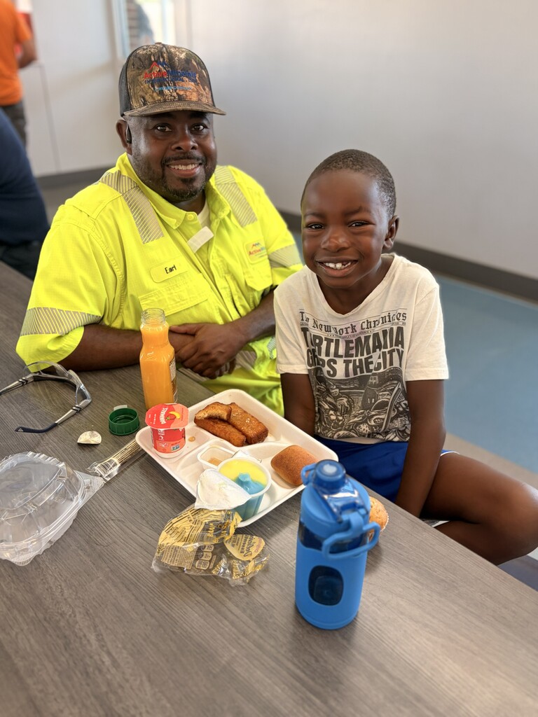 A man in a reflective vest and a young boy smile and sit at a table. A tray of food, drinks, and utensils are on the table.