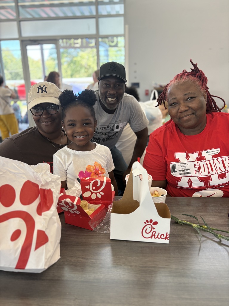 Three people sit at a table with Chick-fil-A bags and boxes, smiling for a photo. A young girl holds a box. Behind them, people are walking, and there are trees outside.