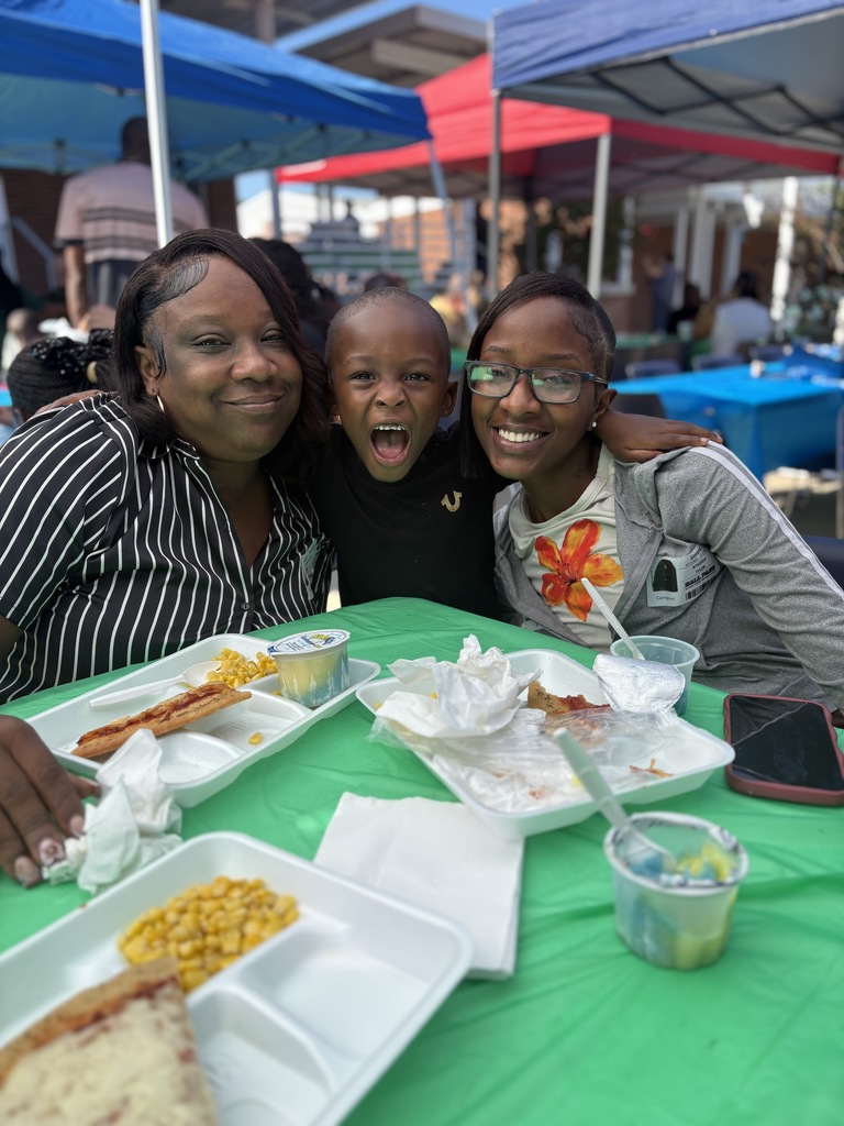 Three people sit at a table with trays of food. One person has an open mouth. A green tent is in the background.