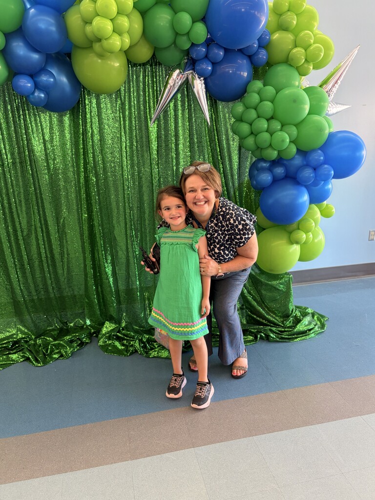 An adult woman and a young girl stand together in front of a green balloon arch.