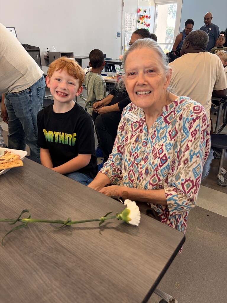 A woman and a boy sit at a table with a white flower in front. People sit behind them.