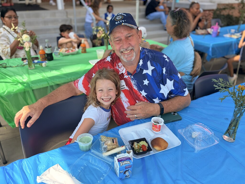 An adult and a child share a table at an outdoor event, smiling and seated at a blue table.