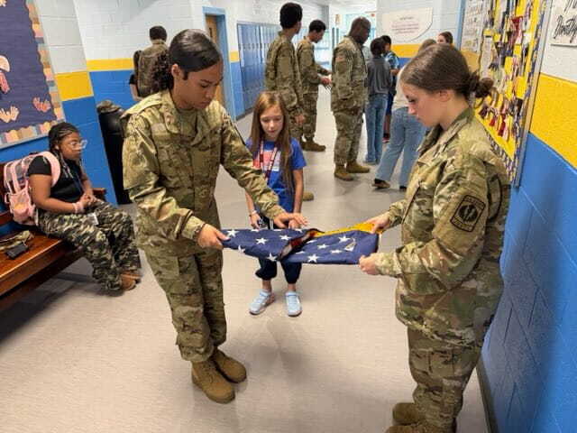 Two women in military uniforms fold a flag while a young girl watches. People are in the background.