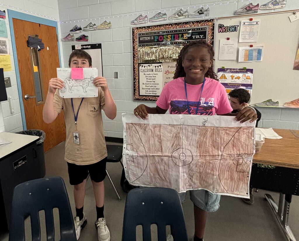 Two children stand in a classroom, each holding a piece of paper. One displays a small, folded paper, and the other holds a larger, unfolded sheet.