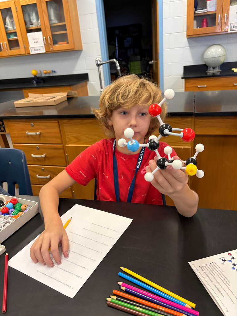 A student in a science lab, seated at a table, holding a molecular model and working on paper.