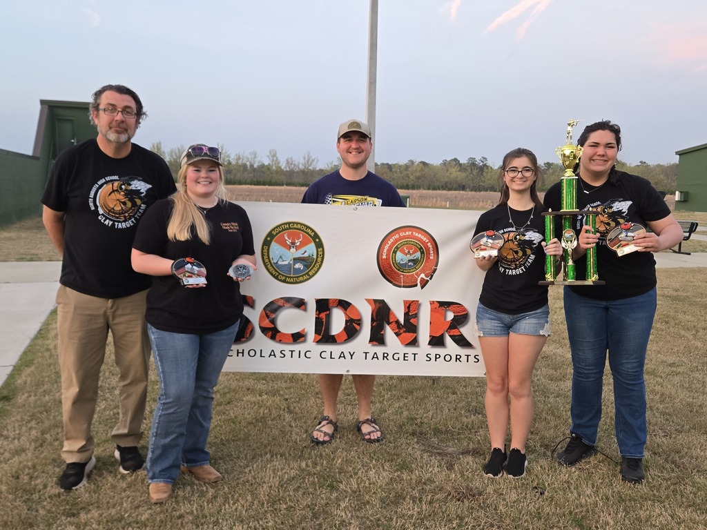 Six people stand behind a banner that reads "SCDNr" and hold trophies. They are all dressed in black.