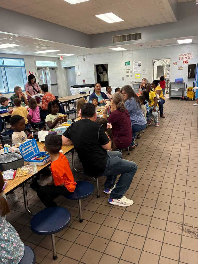 A cafeteria with children and adults seated around tables. They are eating. The floor is tiled and there are glass windows.