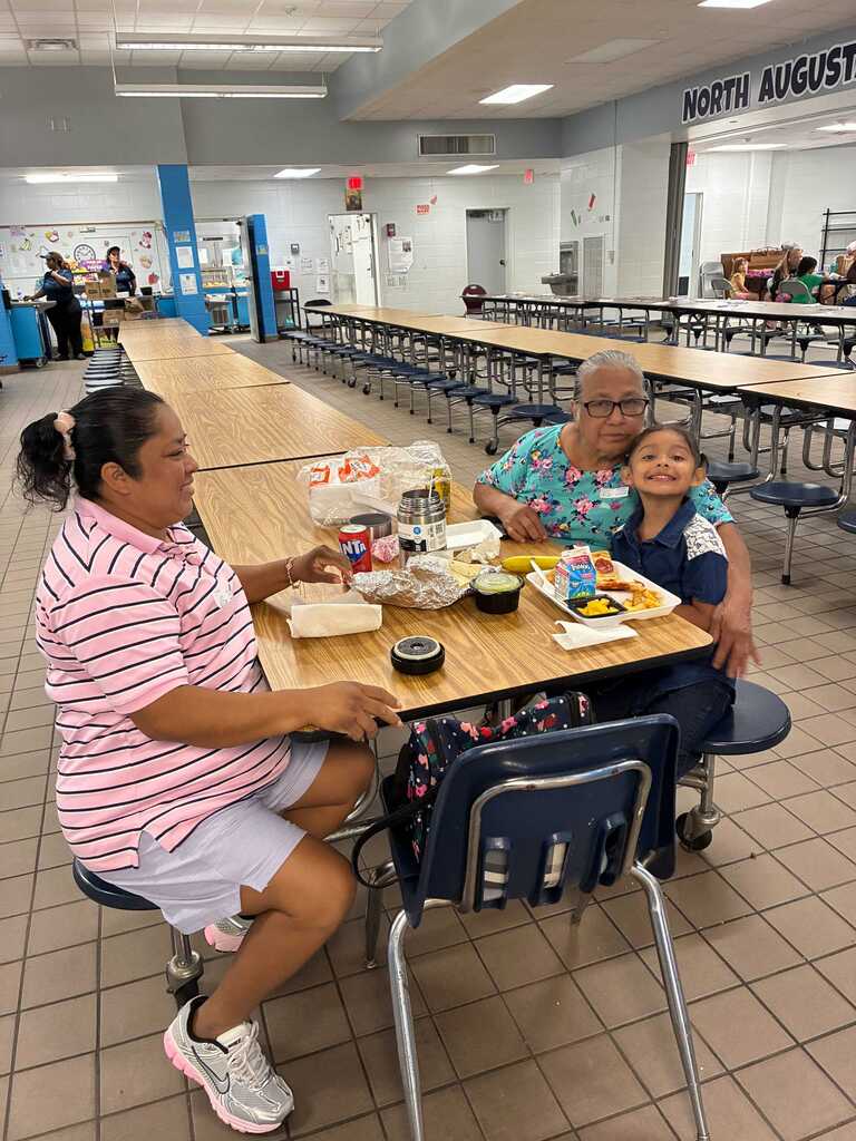 People sit at a cafeteria table with trays of food. A sign in the background reads "North August".