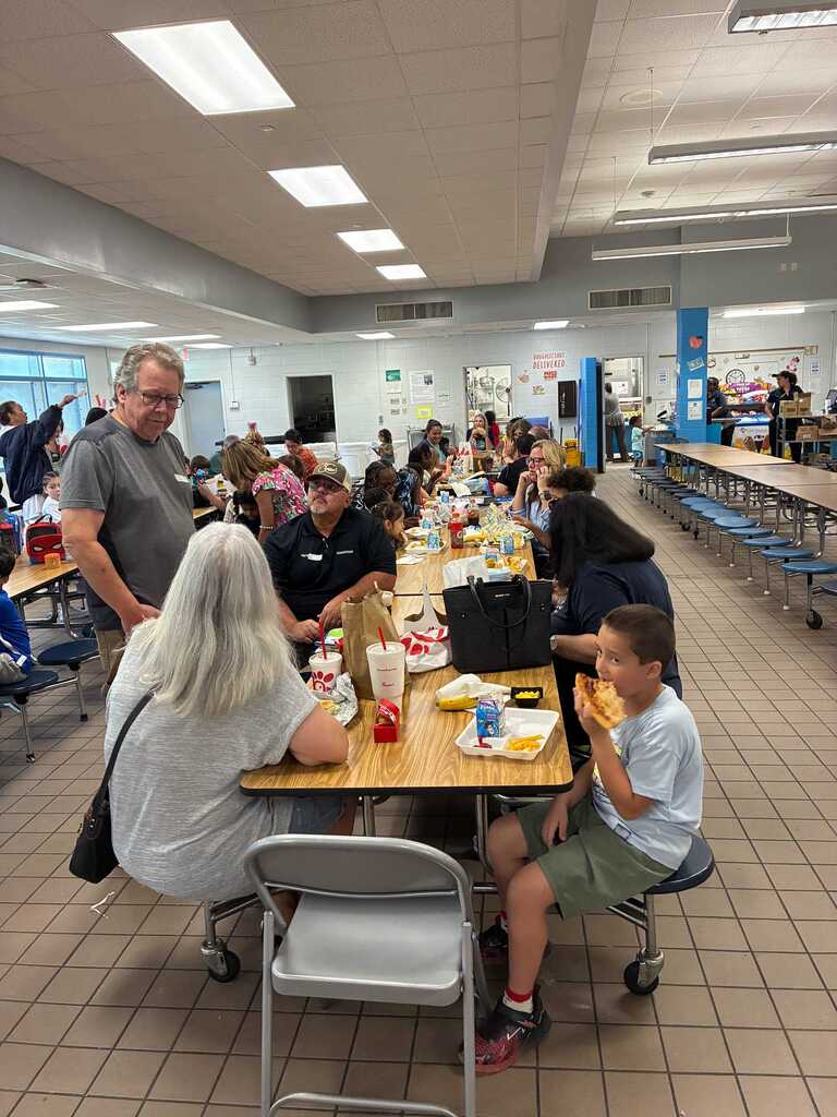 People seated at tables in a cafeteria. Some are eating. The room has a tiled floor and overhead lighting.