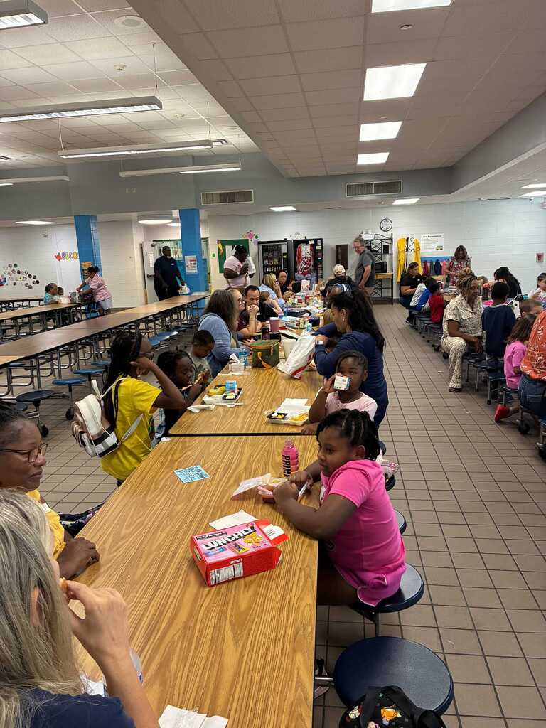 People seated around tables in a cafeteria with some engaged in conversation. A child holds a box of games.