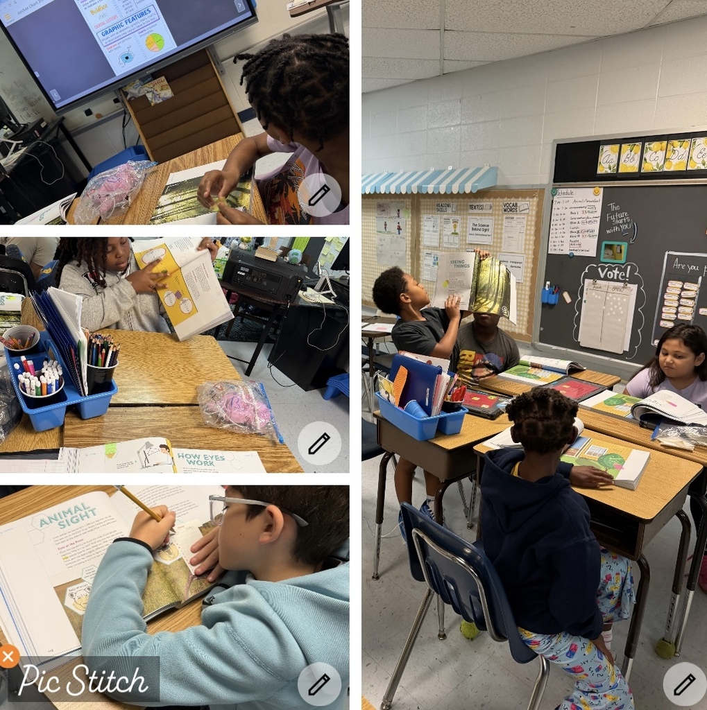 Four students in a classroom with desks, books, and school supplies. One child reading aloud.