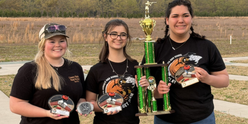 Three women hold trophies and medals; one trophy is gold, while others are silver. They wear black T-shirts.