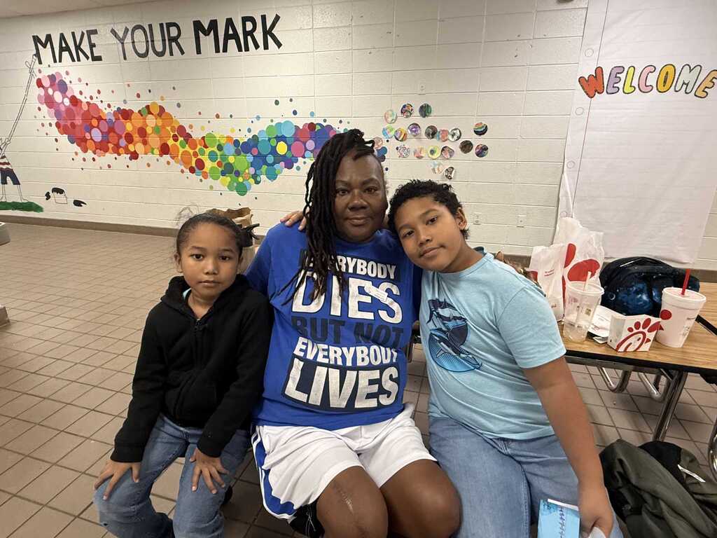 Three individuals seated, one wearing a blue shirt with "Everybody Dies, Everybody Lives" text. Background includes a motivational wall art.