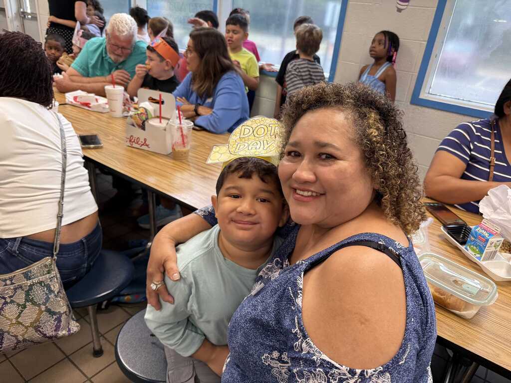 Woman and child sitting at a table in a cafeteria. Other people sit at tables in the background.