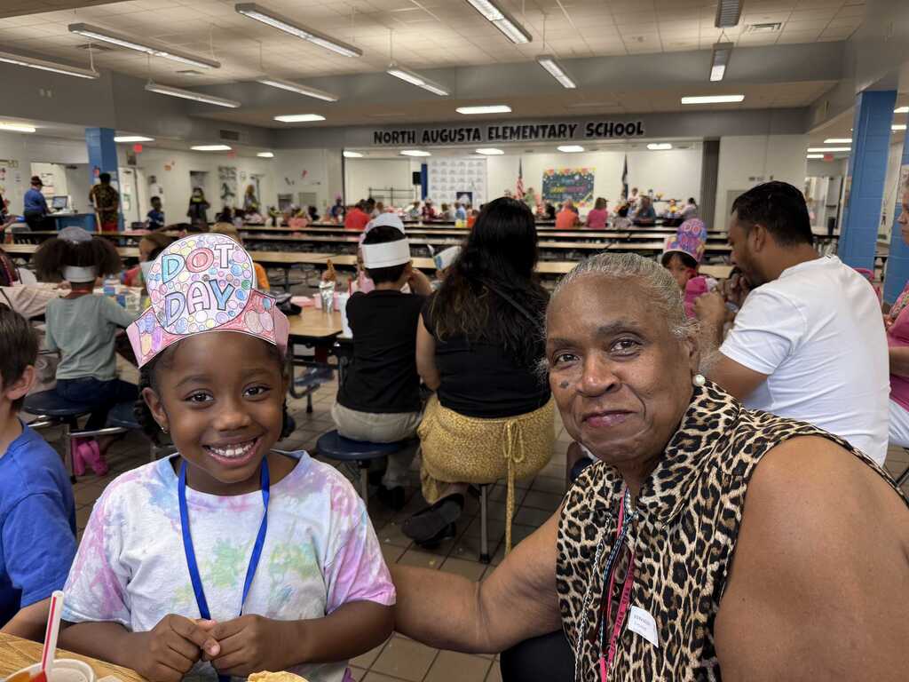 A woman wearing a leopard print shirt and a child in a tie-dye shirt sit together in a cafeteria.