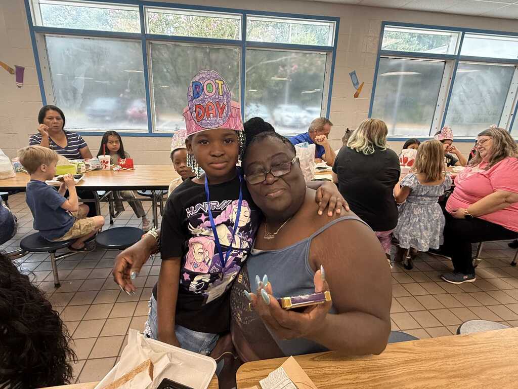 A person with glasses holds a phone, posing with a child in a birthday hat. Behind them, people sit at tables in a cafeteria with large windows.