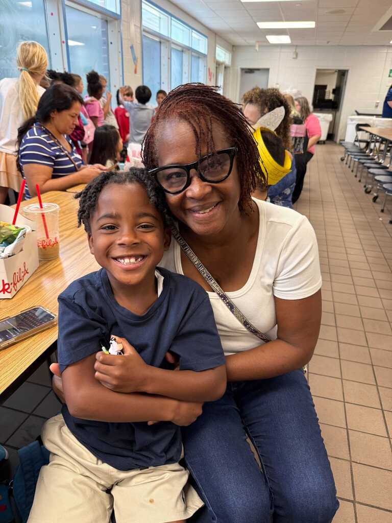 Woman with glasses smiles while sitting with a child who holds a stuffed animal. Other people sit at tables in the background.